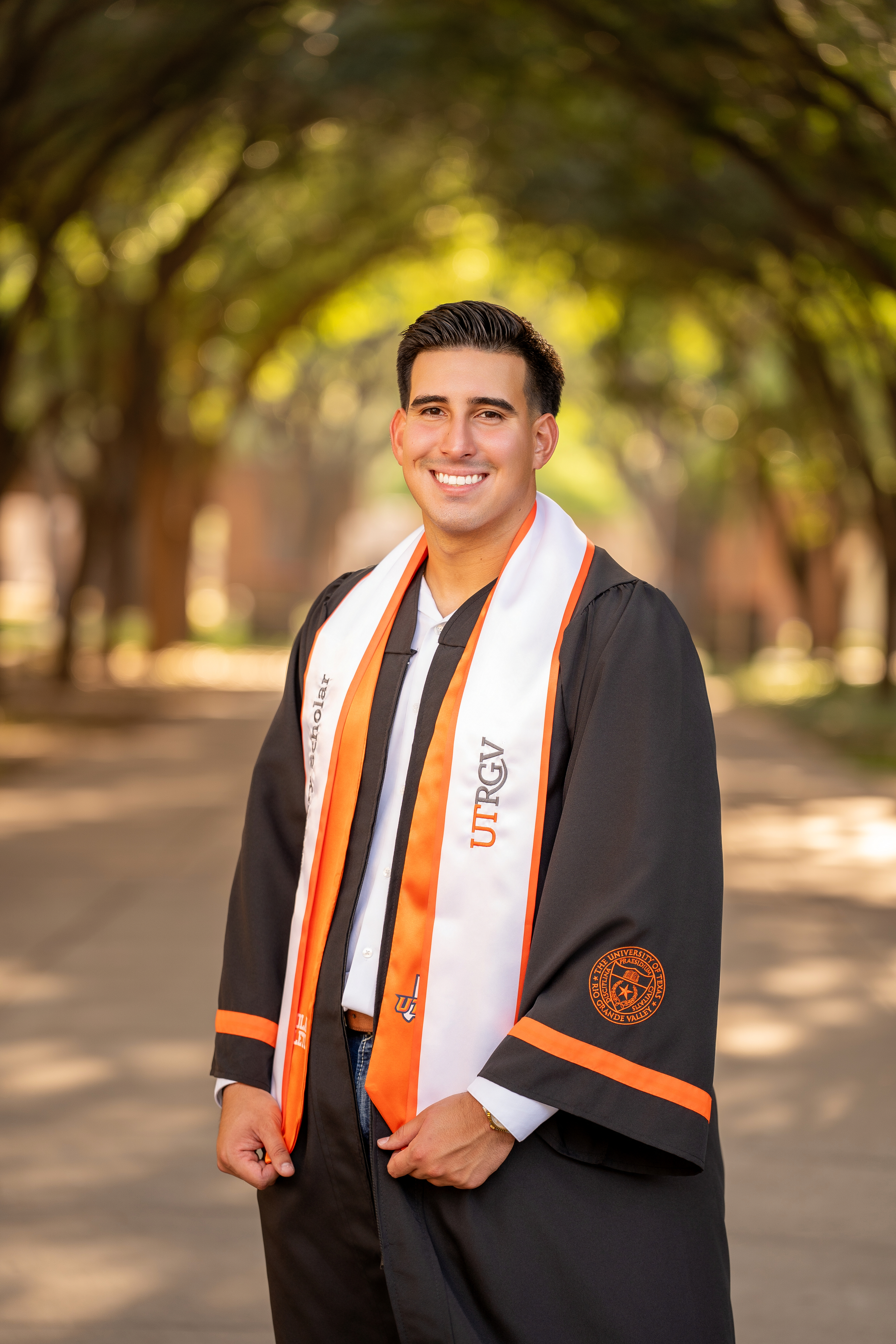 A person wearing a University of Texas Rio Grande Valley graduation gown and a white-and-orange stole stands on a tree-lined campus walkway.