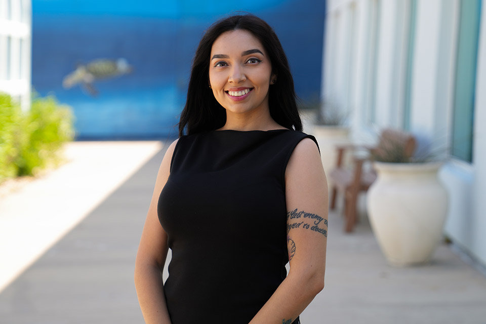A person wearing a sleeveless black dress stands outdoors along a walkway beside a modern building with large windows and potted plants. A blue mural featuring a sea turtle is visible in the blurred background, and a tattoo is visible on the person's upper arm.