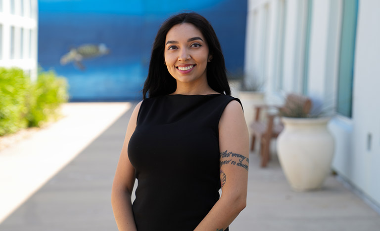 A person wearing a sleeveless black dress stands outdoors along a walkway beside a modern building with large windows and potted plants. A blue mural featuring a sea turtle is visible in the blurred background, and a tattoo is visible on the person's upper arm.