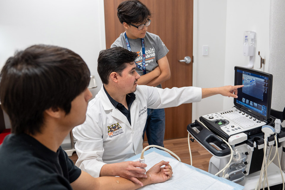Doctor in a white coat points at an ultrasound image on a screen, explaining it to a patient seated nearby. A medical assistant observes attentively.