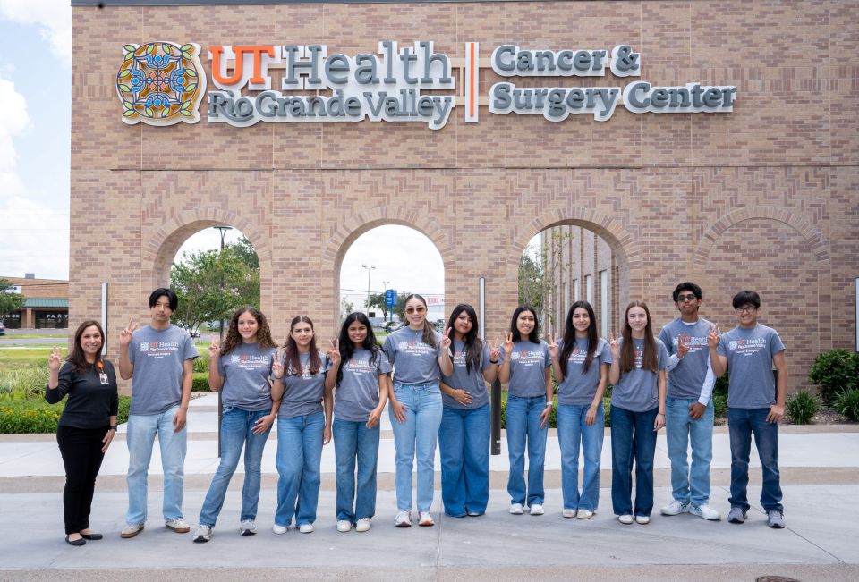A group of young people and adults stand in front of a building with the sign "UT Health Rio Grande Valley Cancer & Surgery Center," posing and making hand signs.