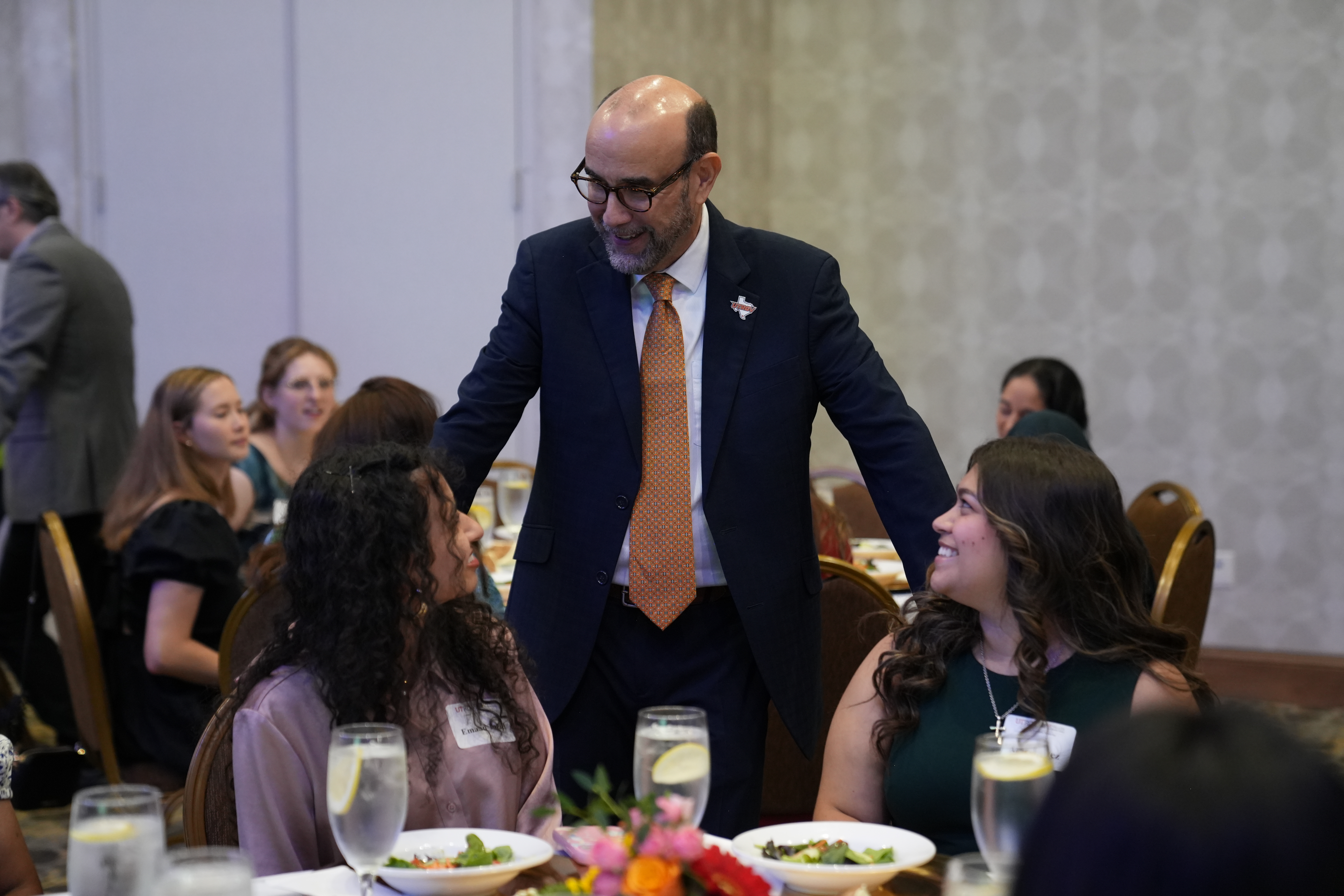 A person in a dark suit and orange tie stands between two round banquet tables, speaking with attendees seated during a formal event. Several guests are seated around the tables with plates of salad and glasses of water, while additional attendees are visible in the background of the conference-style dining room.