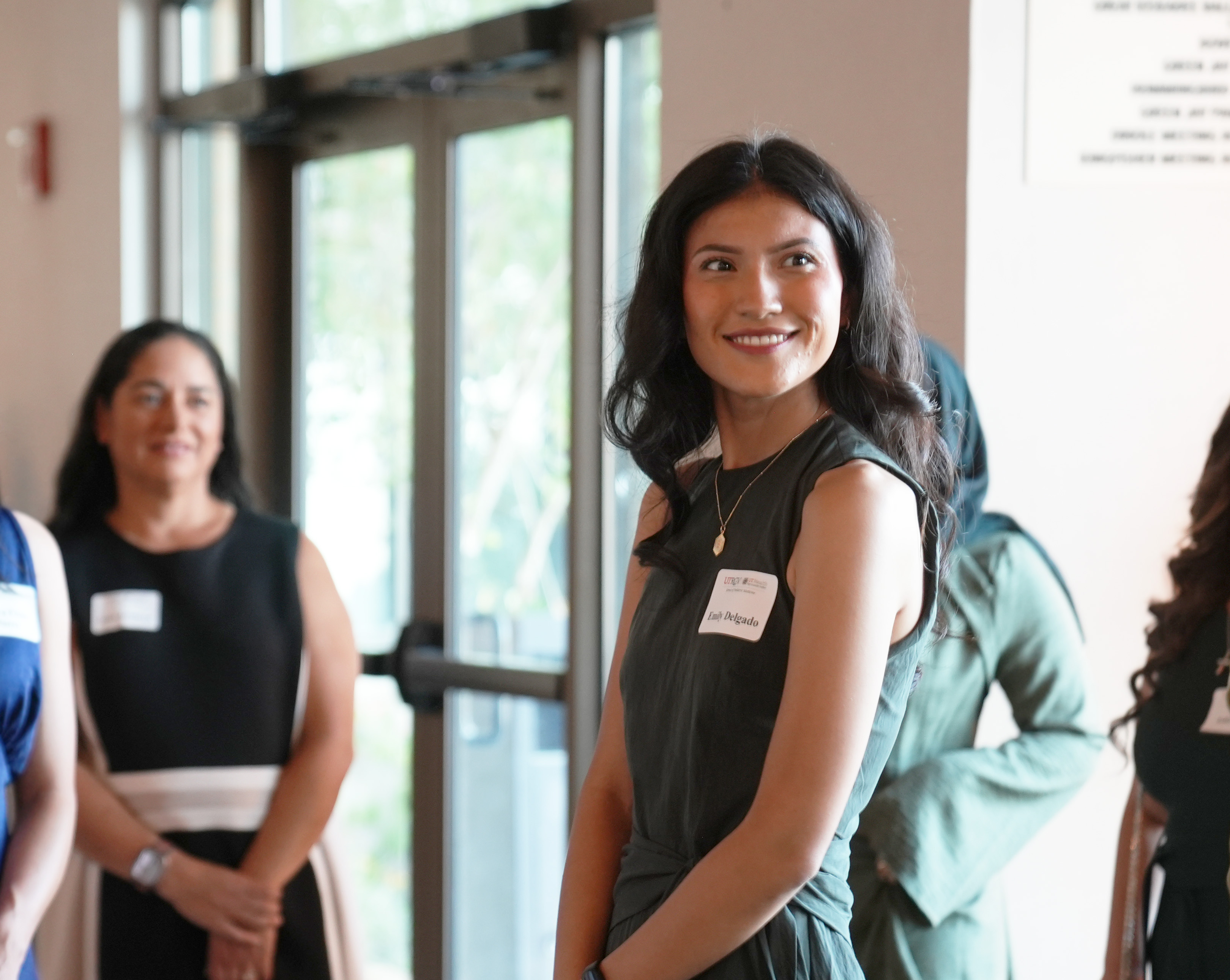 A person wearing a sleeveless dark green outfit and a name tag stands near a glass entrance during a networking or reception event. Several other attendees are visible in the background, and natural light streams through the large windows and doors of the venue.