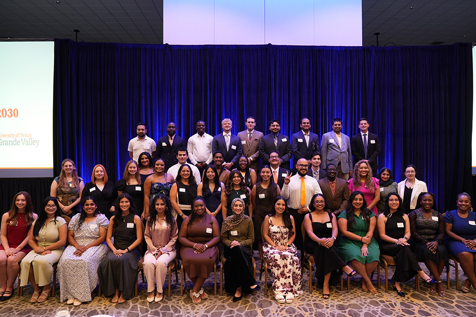 Group photo of students and faculty participants at a university dean's reception, posed on a stage in front of blue curtains during a recognition event.
