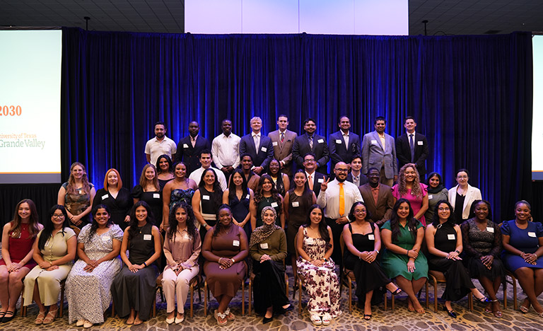 Group photo of students and faculty participants at a university dean's reception, posed on a stage in front of blue curtains during a recognition event.