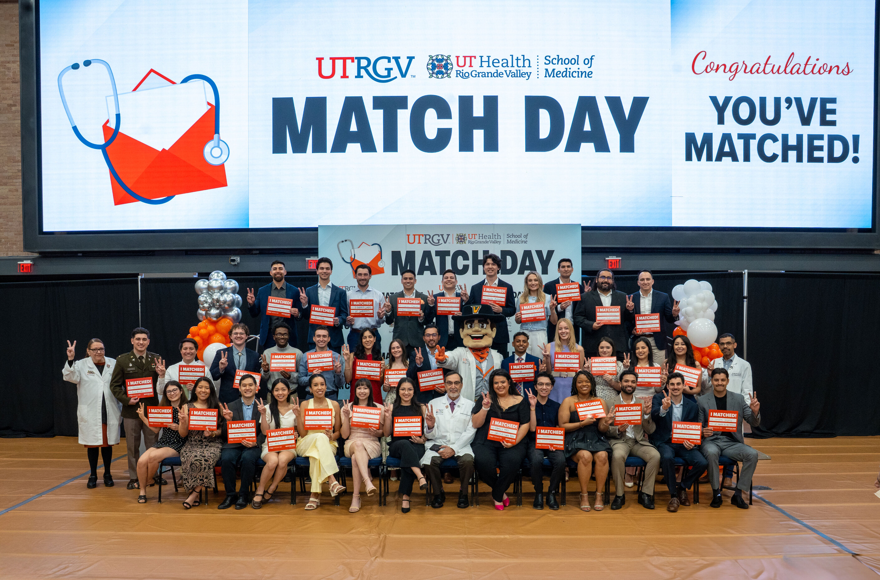 A group of medical students pose indoors, holding red "I MATCHED" signs, in front of a "MATCH DAY" banner at a UTRGV School of Medicine event.