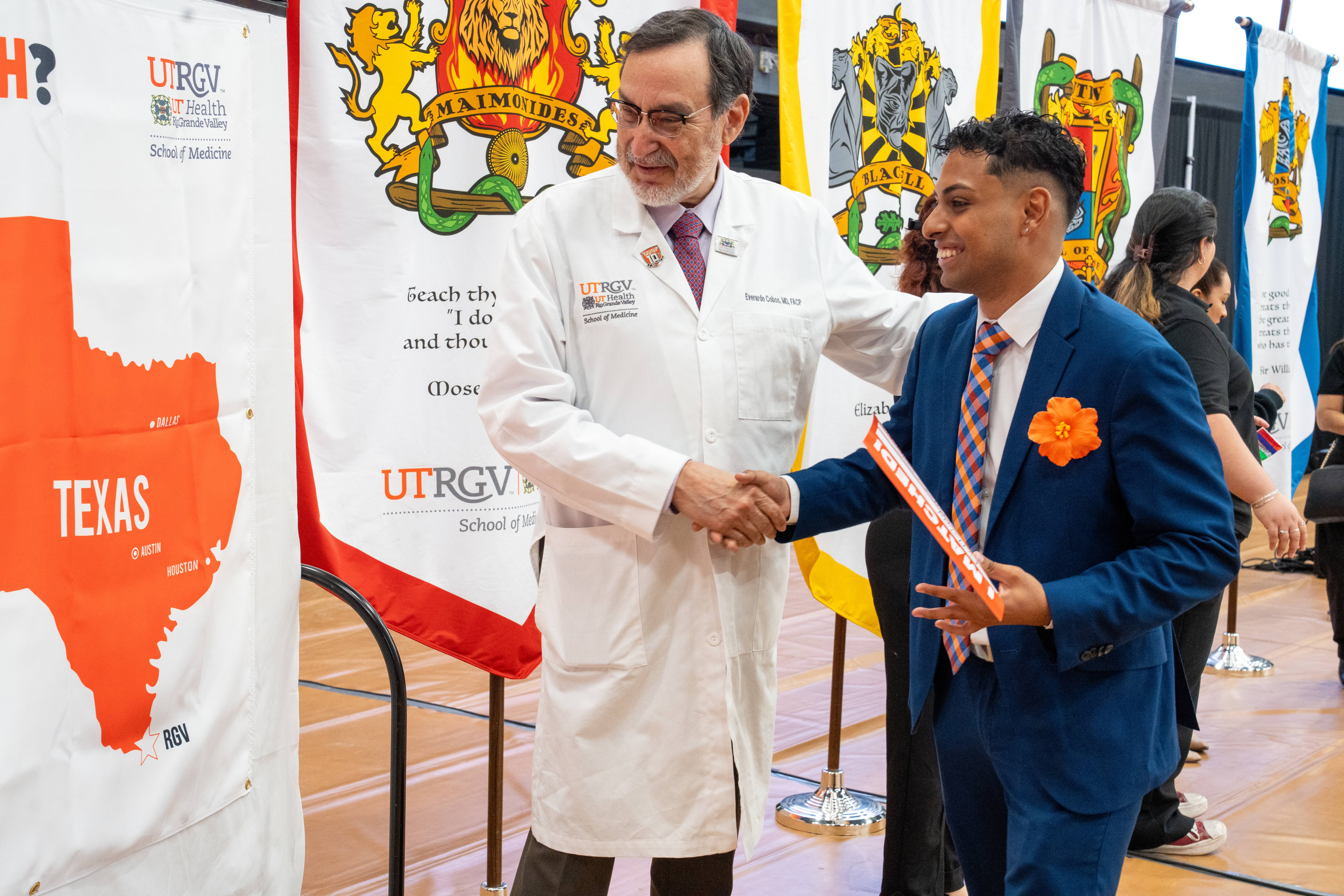 A man in a white coat and a man in a suit shake hands, smiling warmly. Behind them are colorful banners, creating an academic and celebratory tone.