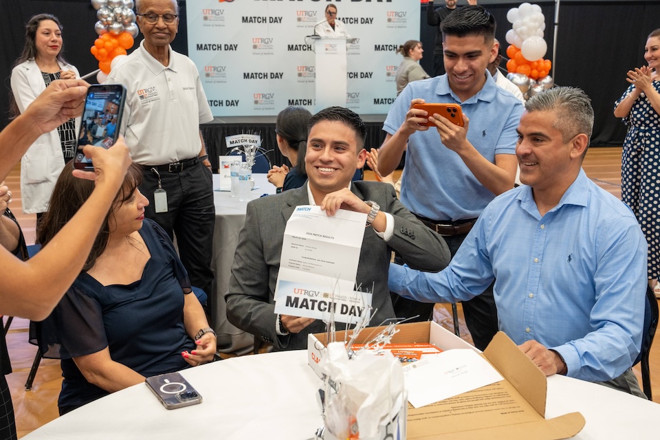 A joyful moment on Match Day as a person holds a paper, surrounded by smiling family and friends. Cameras capture emotions in a festive setting.