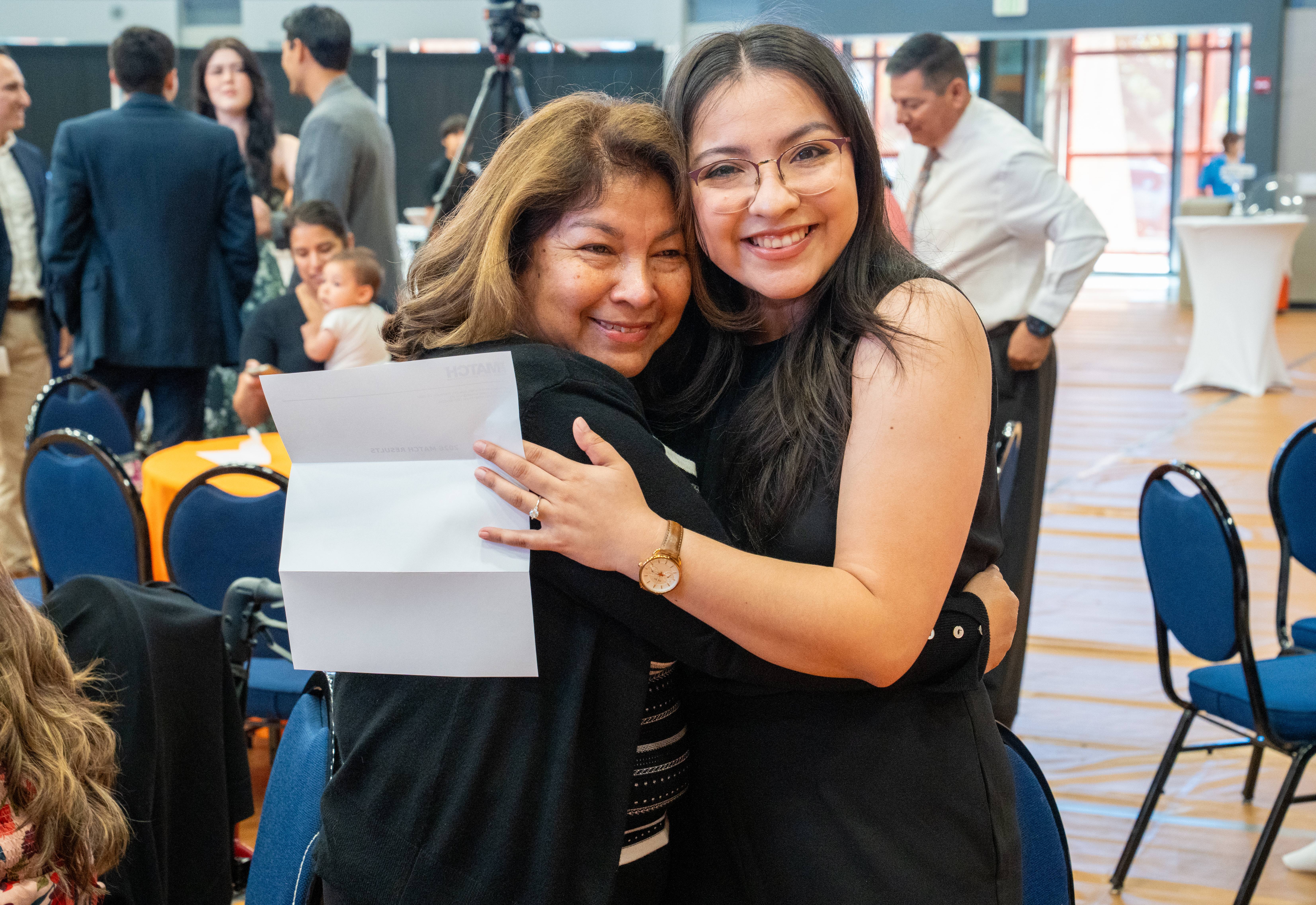 Two women hugging and smiling in a conference room. The woman on the right holds a paper. Chairs and people are visible in the blurred background.
