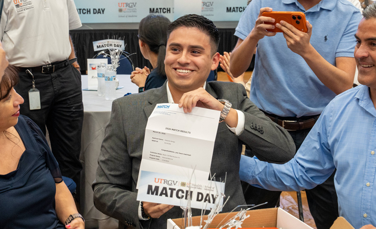 A joyful moment on Match Day as a person holds a paper, surrounded by smiling family and friends. Cameras capture emotions in a festive setting.