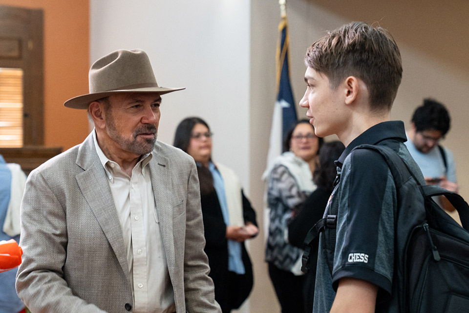 A UTRGV Chess team member wearing a backpack speaks with another person at an indoor gathering.