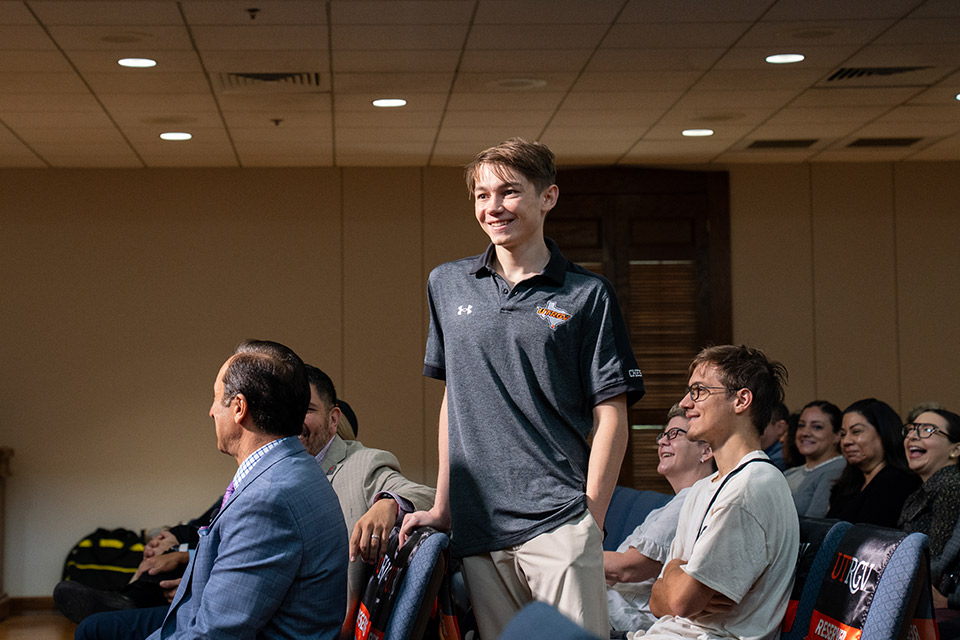 A person wearing a UTRGV Chess polo stands among seated audience members during an indoor event.