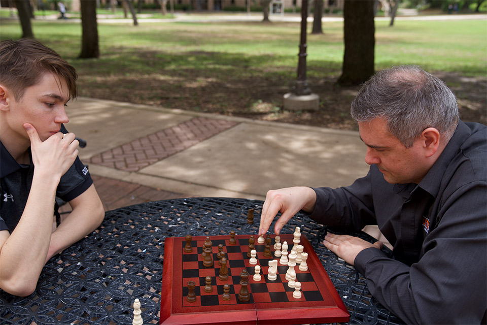 Two people play chess at an outdoor table, with one moving a white chess piece while the other watches the board.