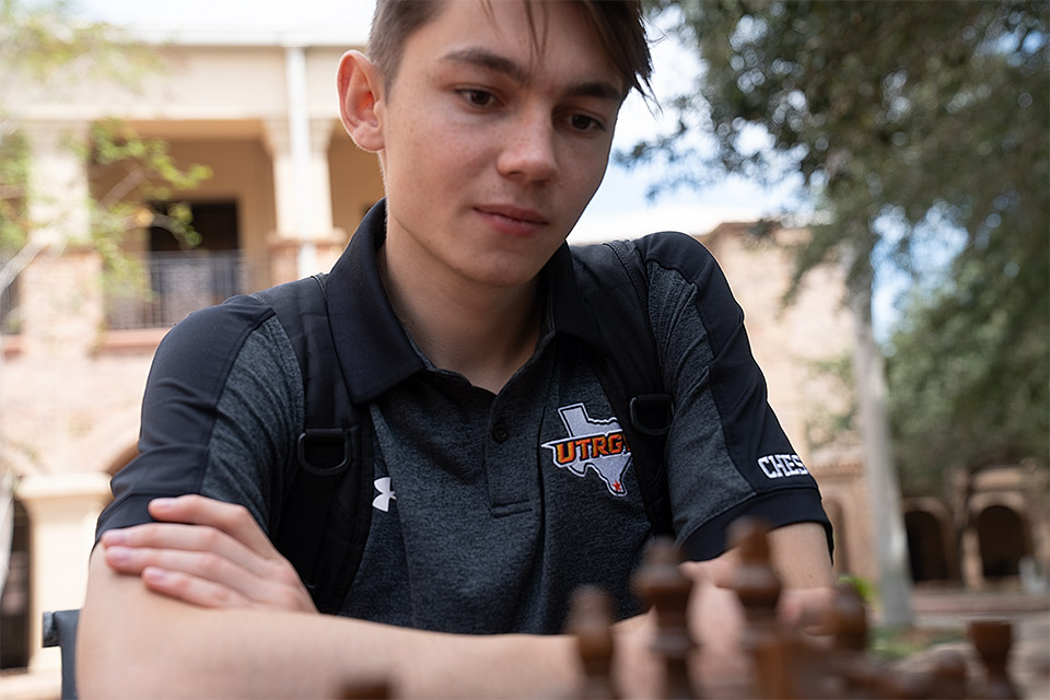 A UTRGV Chess team member looks down at a chessboard while seated outdoors on campus.