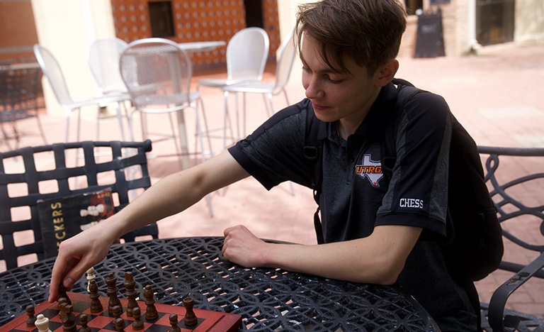 A UTRGV chess player moves a chess piece on an outdoor table while wearing a UTRGV Chess polo.