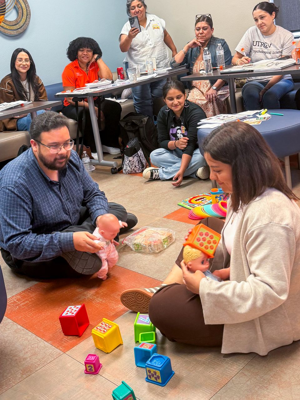 Participants in a UTRGV training session sit in a circle observing two individuals on the floor using dolls and children's toys to demonstrate child-focused interaction techniques.