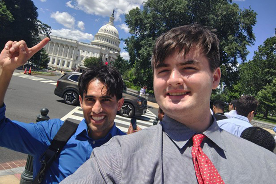 Two students take a selfie near the U.S. Capitol in Washington, D.C., with one pointing toward the building.