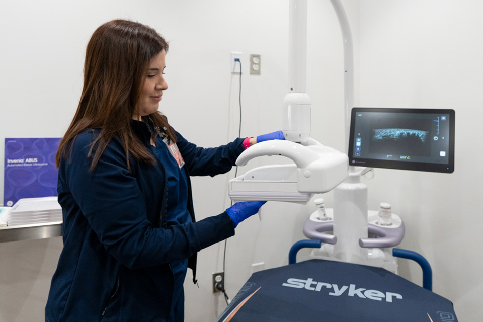 A woman wearing blue gloves adjusts a breast ultrasound machine beside a monitor displaying an ultrasound image.
