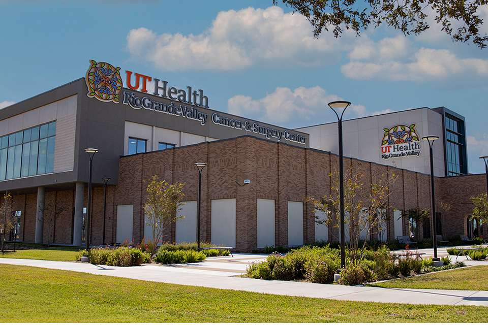 Modern brown brick building labeled "UT Health Rio Grande Valley Cancer & Surgery Center" surrounded by landscaped grounds under a blue sky.