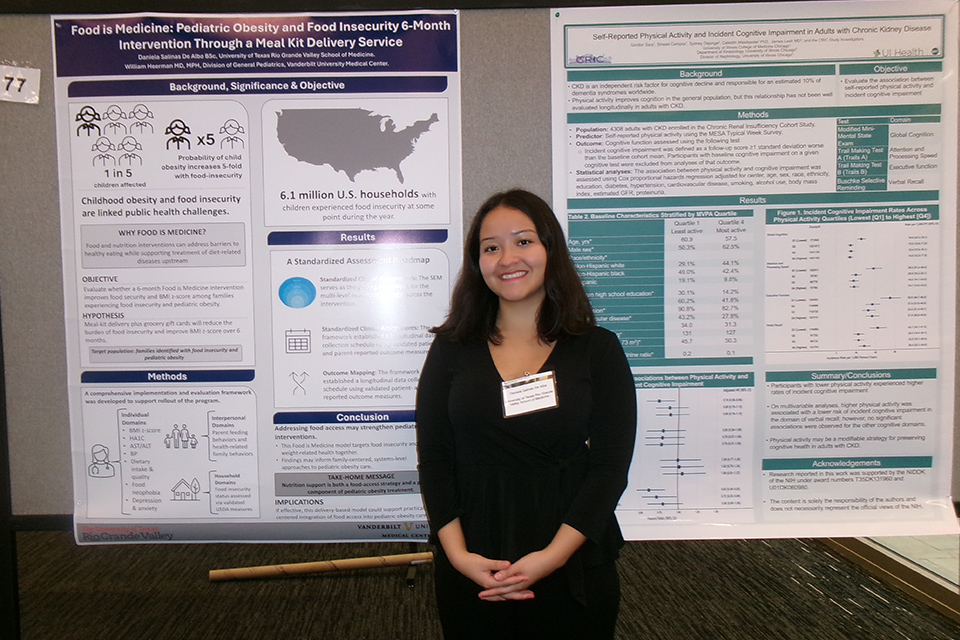 Daniela Salinas De Alba stands in front of research posters on pediatric obesity and food insecurity, and the relationship between physical activity and cognitive impairment in adults with chronic kidney disease.