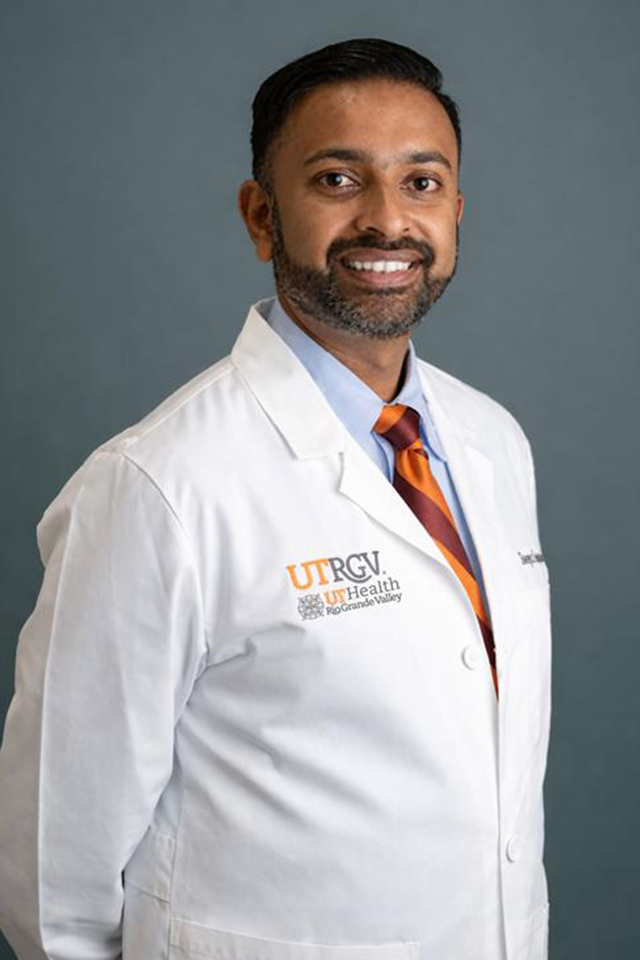 A confident man in a white lab coat with the UTRGV logo, wearing an orange-striped tie, stands against a gray background, smiling warmly.