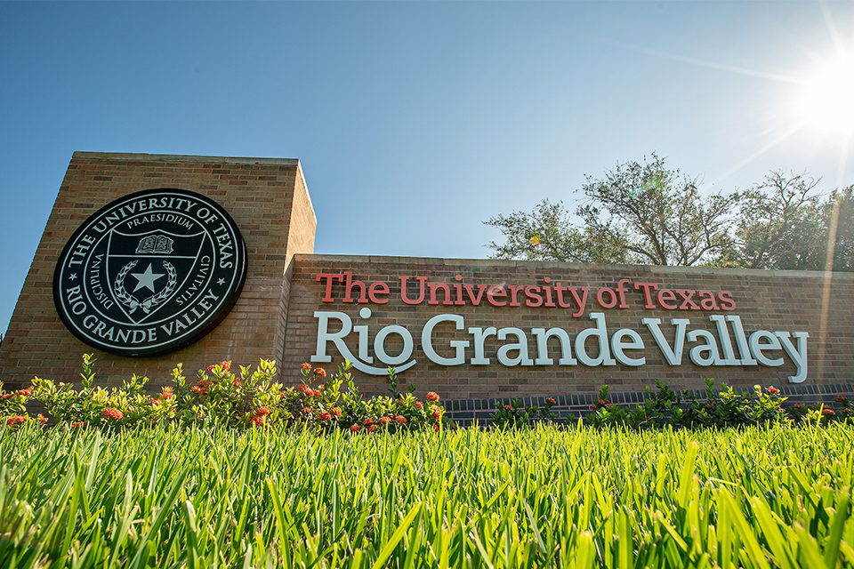 Low-angle view of The University of Texas Rio Grande Valley entrance sign, surrounded by grass and flowers under a sunny blue sky.