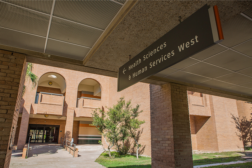 Exterior entrance to the UTRGV Health Sciences and Human Services West building, with an overhead sign directing visitors to the facility.