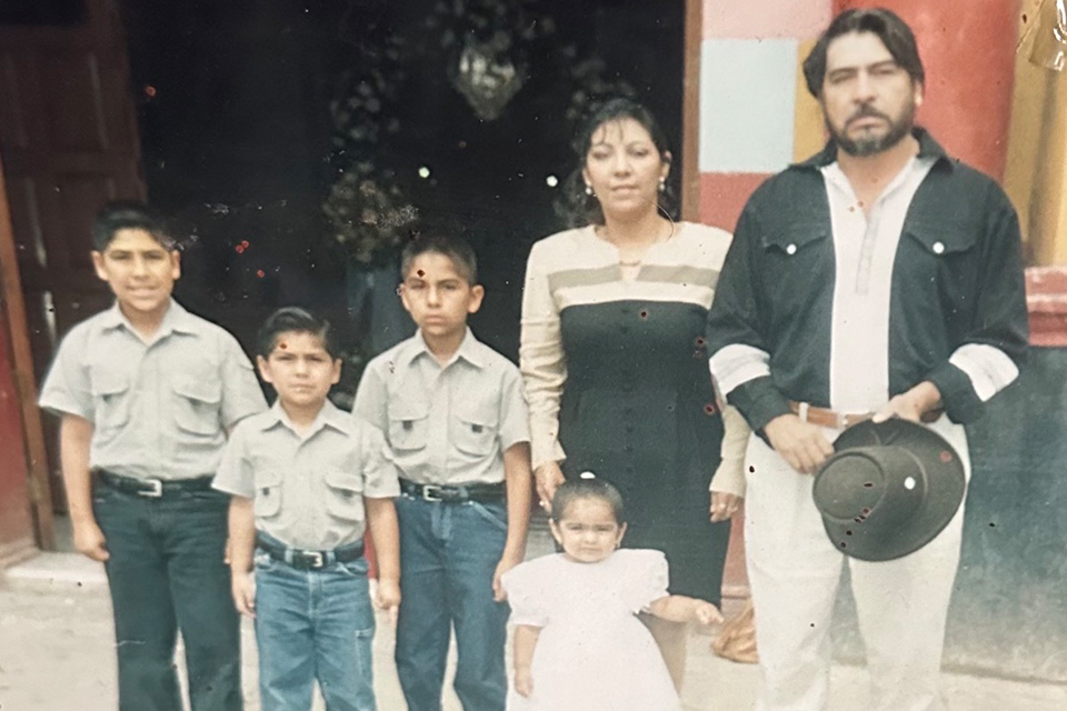 A family of six poses for a photo outside a building. Three boys wear matching shirts, a girl stands in front in a white dress, and two adults stand behind them. The man holds a hat.