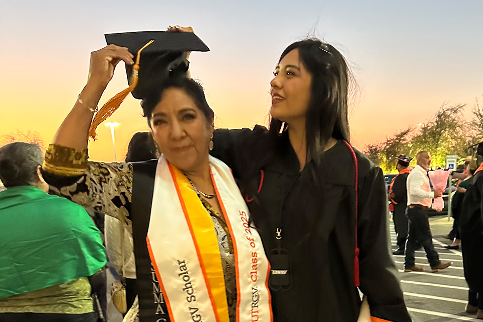 Two individuals are shown outdoors at a graduation ceremony; the person in the foreground, wearing a cap and gown with a sash that reads "UTRGV Scholar Class of 2025," is holding a graduation cap.