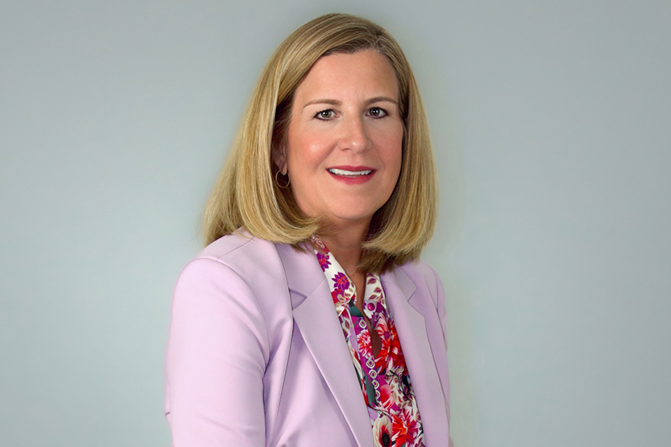 Dr. Magdalena Hinojosa smiles for a professional headshot, wearing a light purple blazer over a floral blouse against a gray background.