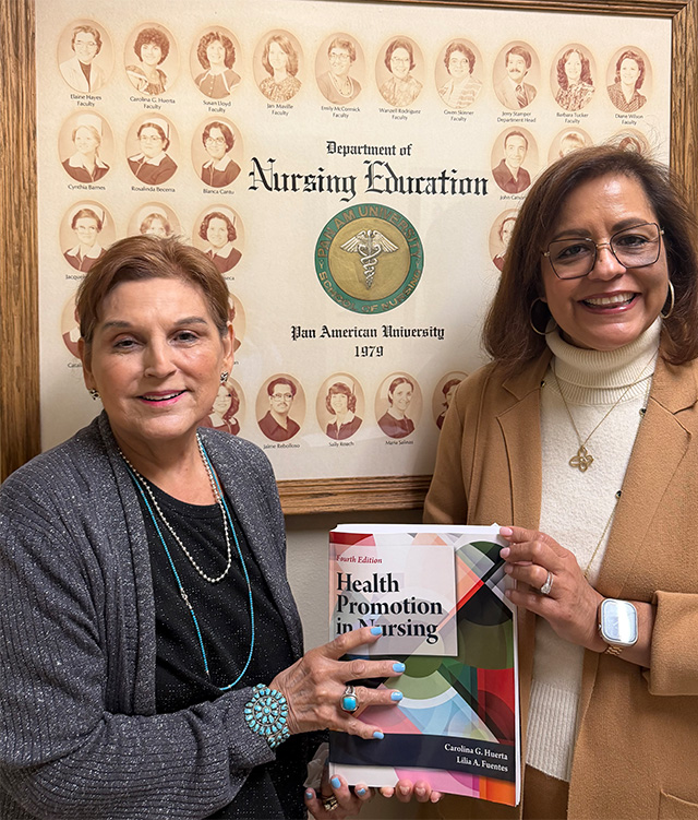 Two females stand next to each other holding a book in the middle of the photo. They stand in front of a wall that include a picture frame.