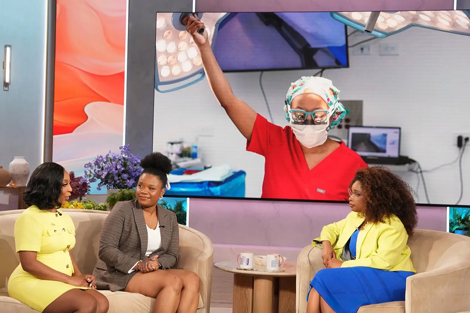 A talk show set features two women in conversation on beige chairs, one in a yellow outfit, the other in gray. Behind them is a large screen showing a surgeon in red scrubs and a mask, raising an arm in an operating room. The atmosphere is professional and engaging. 