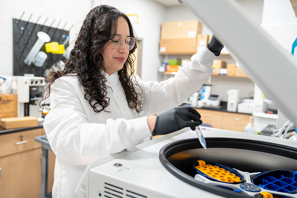 A laboratory technician with long hair and safety glasses works with a centrifuge, wearing a white coat and black gloves, focused and professional. 
