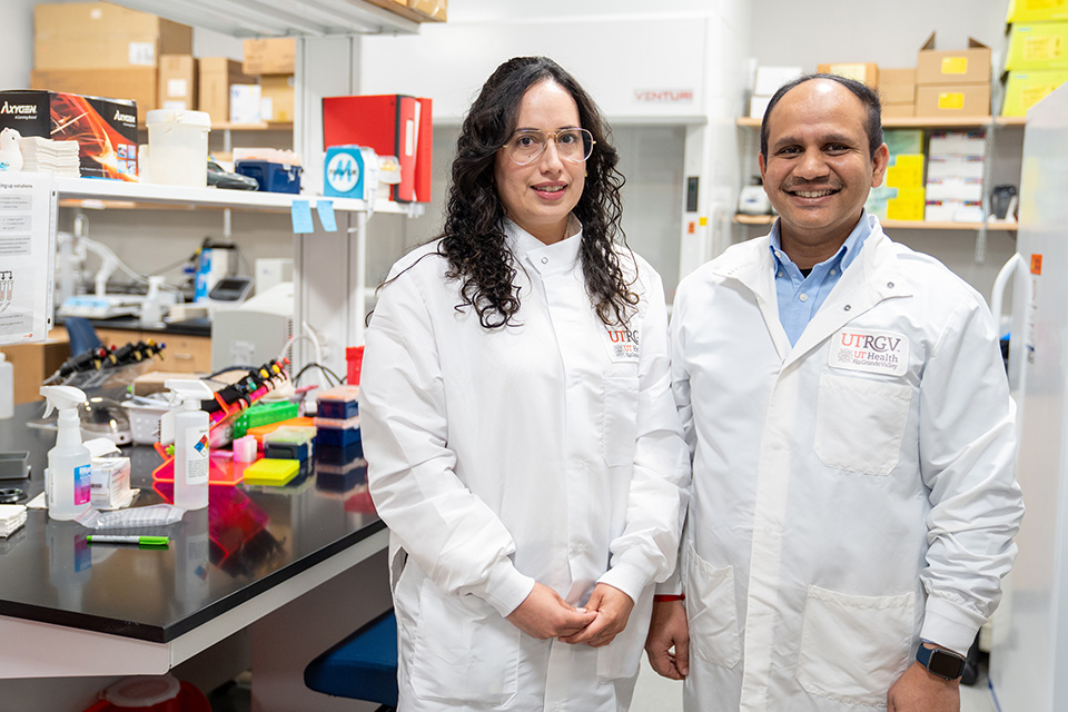 Two lab technicians in white coats stand smiling in a bright, organized laboratory filled with scientific equipment and supplies on shelves.