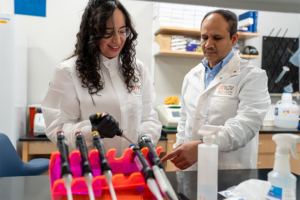 Two researchers in lab coats work in a laboratory. One holds a pipette above a rack, while the other points, suggesting collaboration and focus. 