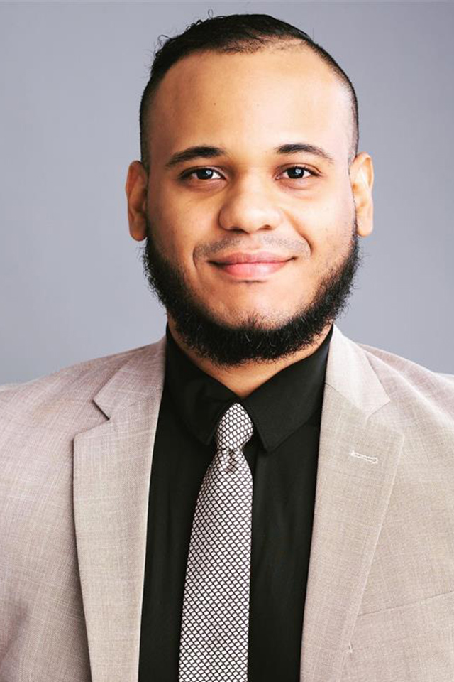 A person wearing a light tan blazer, black dress shirt, and patterned gray tie poses against a plain gray background.  