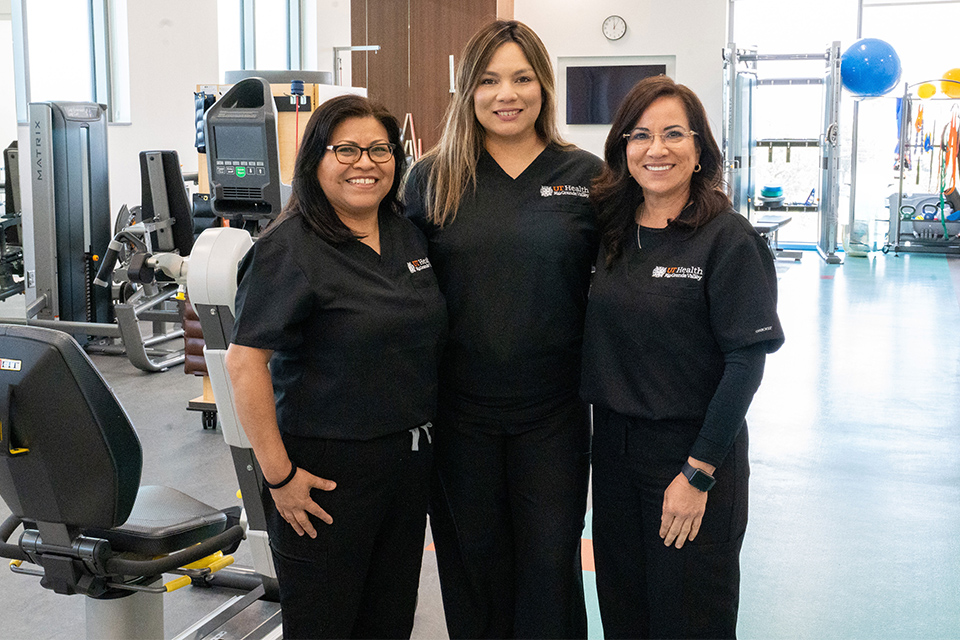 Three smiling women in matching black uniforms with UTRGV logo stand together in a bright, modern gym with exercise equipment, conveying teamwork and friendliness.