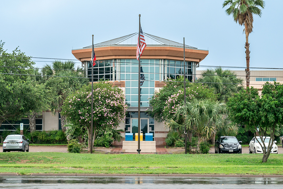 Exterior view of a two-story university building featuring a circular glass rotunda entrance framed by flagpoles, palm trees, and flowering pink crepe myrtles.