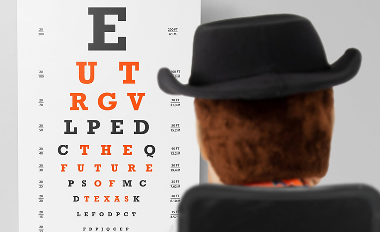 A Snellen eye chart displayed against a light gray background, viewed over the back of a brown plush figure wearing a black cowboy hat. The chart features rows of letters in varying sizes, with key letters highlighted in bold orange that spell out a hidden phrase row-by-row: "UTRGV THE FUTURE OF TEXAS". The remaining letters and measurement ratios appear in standard black text.