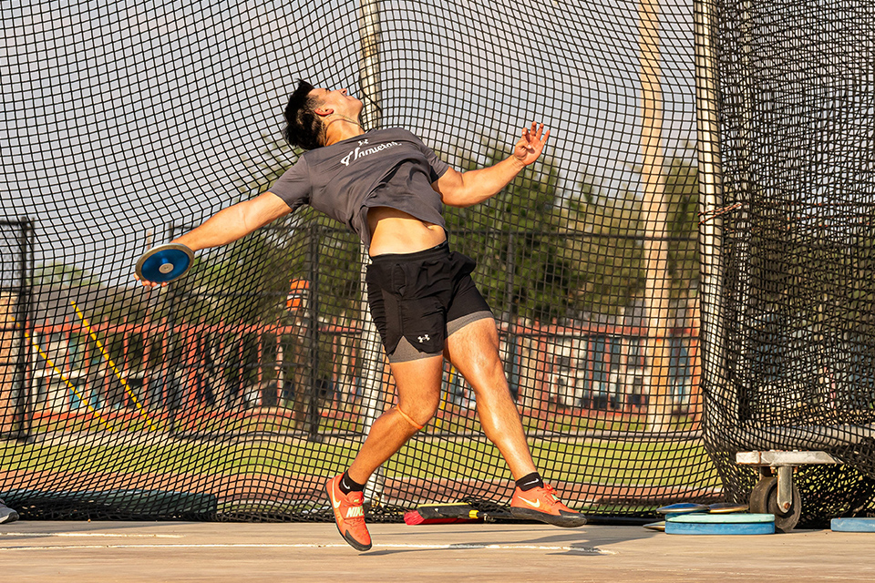 Discus thrower in gray shirt and black shorts launches a blue discus inside a large, netted cage.