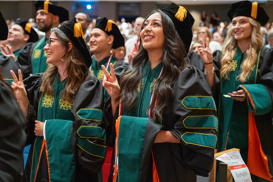 Photo of medical school graduates seated during commencement, wearing black academic regalia with green and gold doctoral hoods. Graduates are smiling and making peace signs as they celebrate the ceremony.