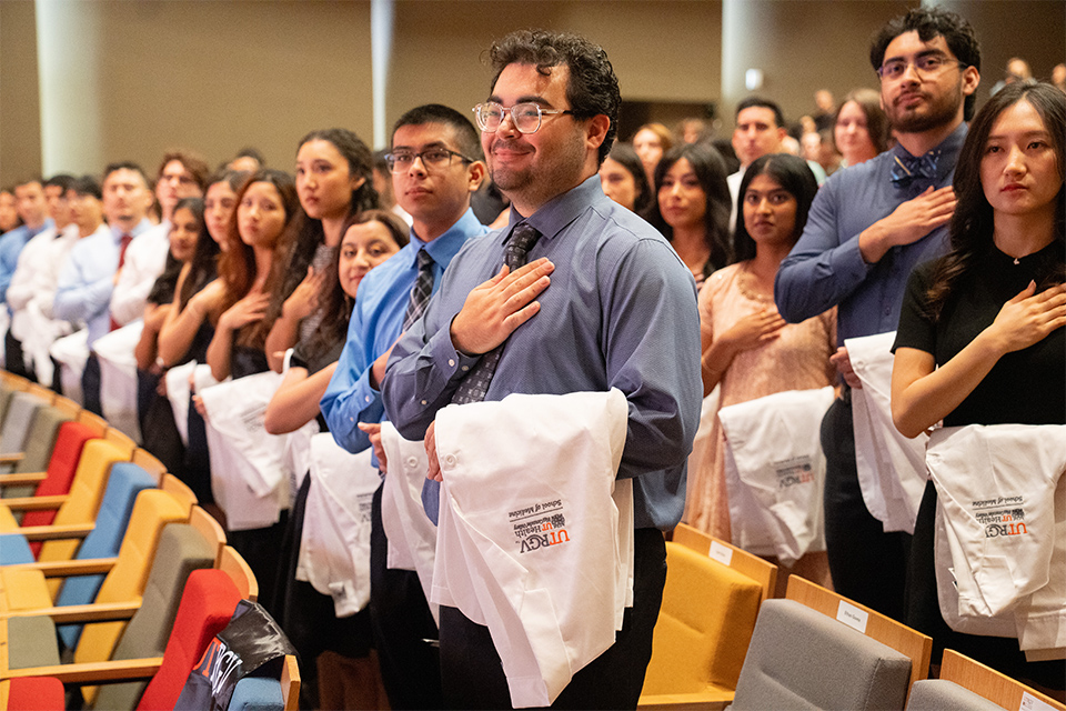 UTRGV School of Medicine students hold white coats and stand with their hands over their hearts during a ceremony.