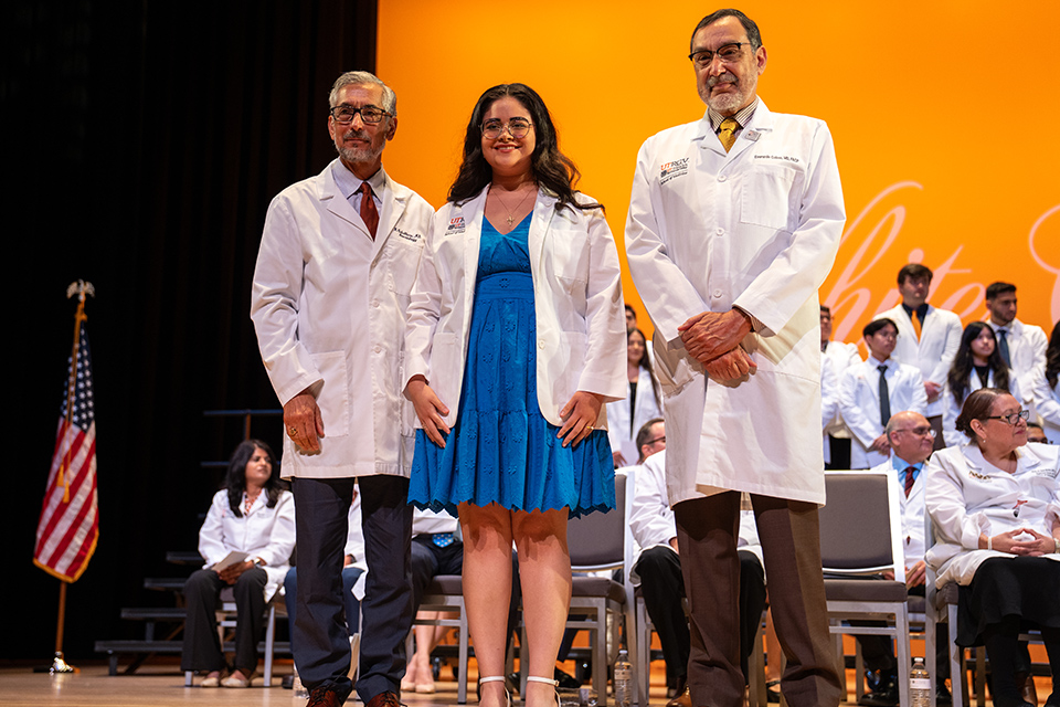 A medical student in a white coat and blue dress stands between two faculty members during a UTRGV School of Medicine ceremony. Other white-coated participants sit and stand onstage behind them.