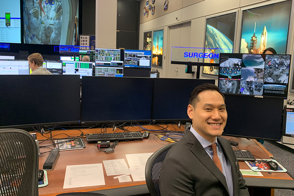 A person in a suit smiles while seated at a desk in a busy mission control room. Multiple monitors display space missions and controls, conveying focus and high-tech activity.