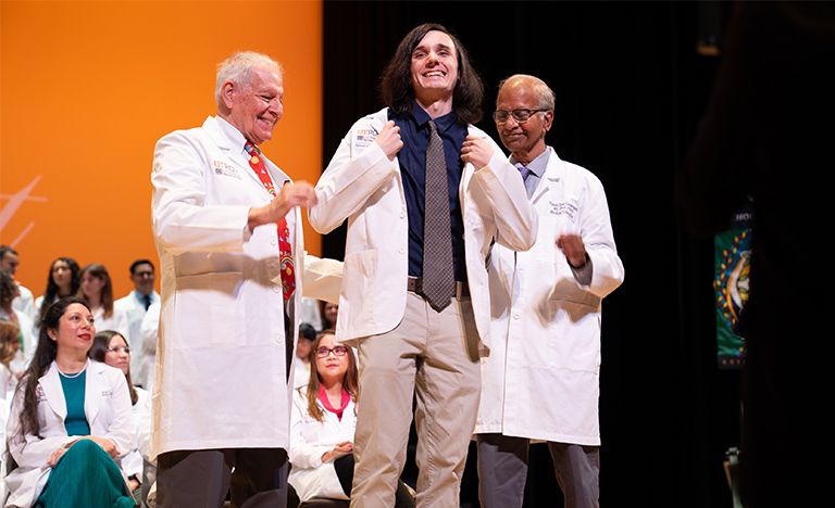 Members of the UTRGV School of Medicine's Class of 2030 celebrate during the annual White Coat Ceremony.  A male smiles big as he puts on his white coat. Two men assist with the coat.