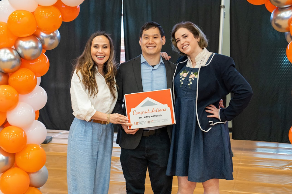 Three people celebrate at an event, smiling and holding a "Congratulations" sign. They stand between festive orange, white, and silver balloons.