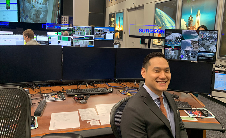 A person in a suit smiles while seated at a desk in a busy mission control room. Multiple monitors display space missions and controls, conveying focus and high-tech activity.