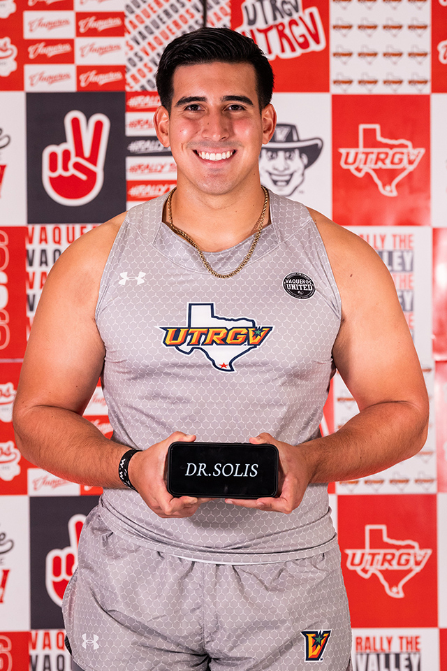 A male athlete in a gray UTRGV uniform stands smiling and holding a plaque that reads "DR. SOLIS" against a red and white UTRGV-themed backdrop.