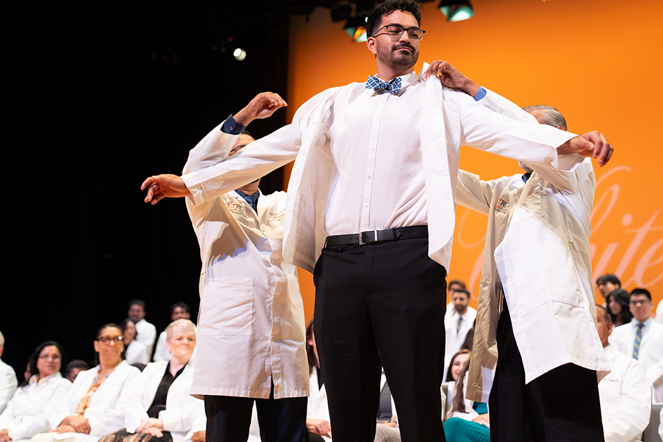 Members of the UTRGV School of Medicine's Class of 2030 celebrate during the annual White Coat Ceremony.  A male smiles big as he puts on his white coat. Two men assist with the coat.