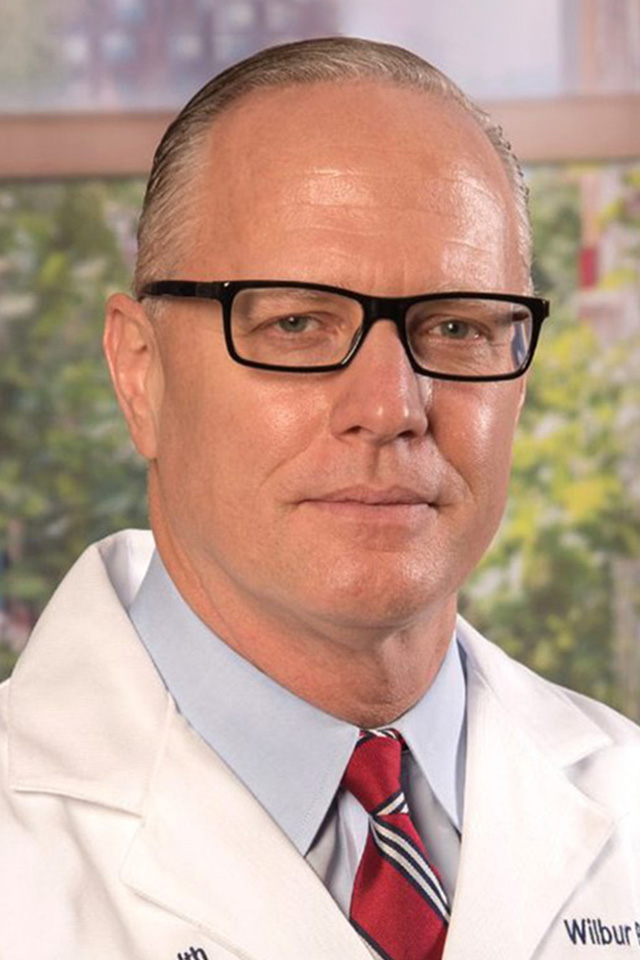 A man wearing glasses, a white lab coat, a blue shirt, and a red striped tie, standing in front of a blurred outdoor background.
